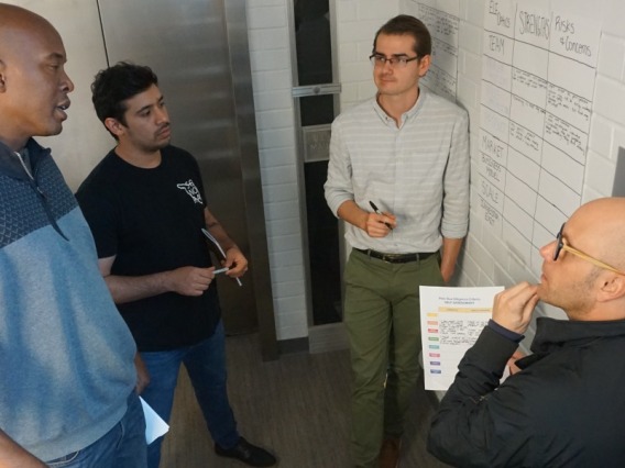Four men talking to each other, standing near a whiteboard with writing on it
