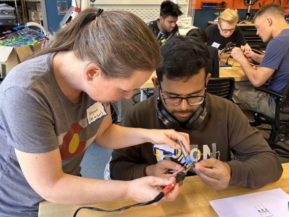 Female professor teaching a male student soldering, with three other students working at another table in the background
