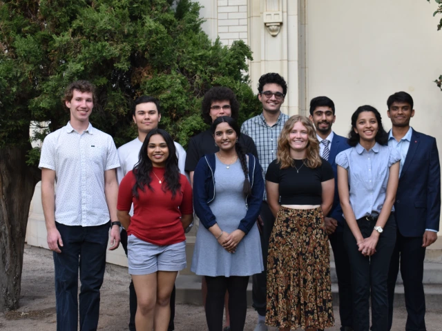 The ten student participants are seen standing outdoors in two rows in front of the white base of the telescope dome in back of Steward Observatory