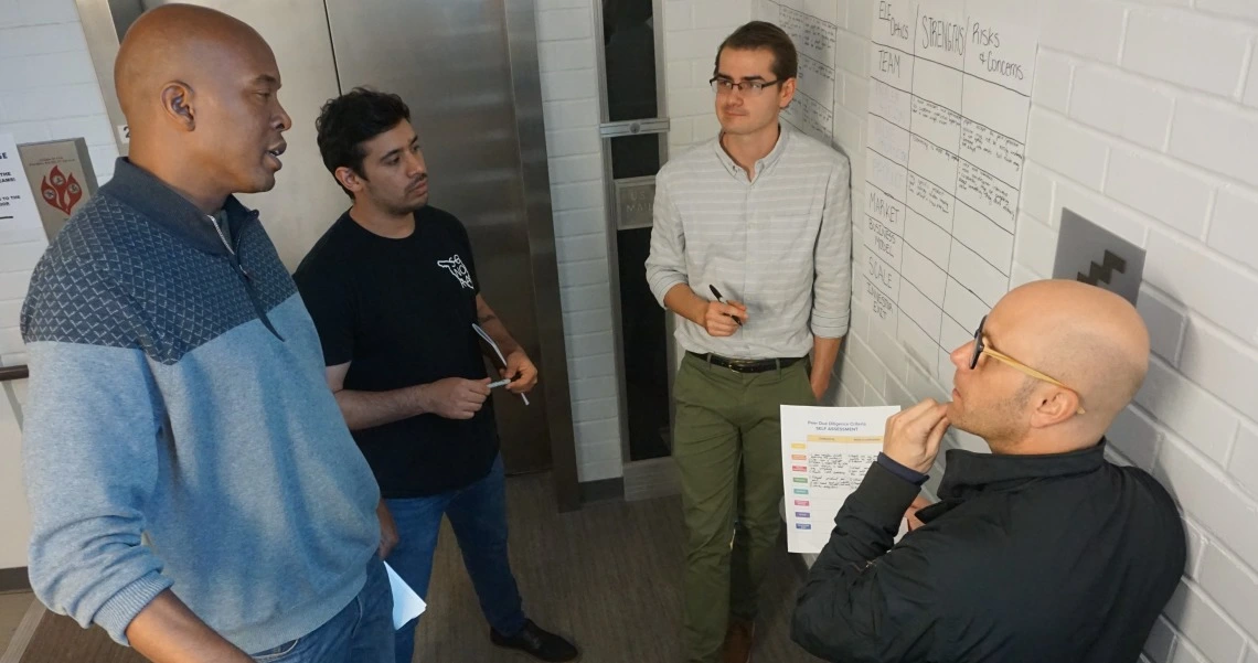 Four men talking to each other, standing near a whiteboard with writing on it