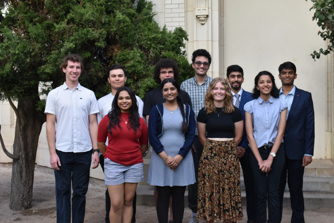 The ten student participants are seen standing outdoors in two rows in front of the white base of the telescope dome in back of Steward Observatory